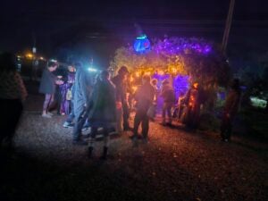 A group of people gather at night in front of a small, decorated structure lit with purple and yellow lights.