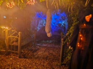 A spooky Halloween scene unfolds like a gallery of frights, featuring a misty path surrounded by dark foliage. A hooded figure looms on the right, while a ghostly decoration floats in the center. Twinkling lights and cobwebs complete this eerie canvas.