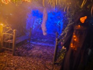A spooky Halloween scene transforms the gallery, featuring a hooded figure holding a carved pumpkin under dim, orange and blue lighting. A cobweb-filled tunnel with a large, wrapped cocoon at its center is visible, lined by foliage and a metal bench on a gravel path.