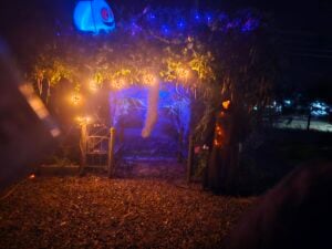 A spooky outdoor Halloween gallery at night showcases a ghostly figure in a dark cloak, cobwebs, and dim orange and blue lights. A glowing jack-o'-lantern and other decorations add to the eerie atmosphere.
