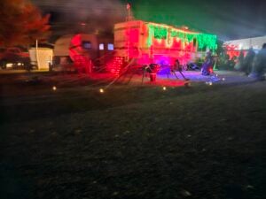 A festive Halloween scene unfolds like a gallery, with a camper adorned in bright red and green lights, spider decorations, and webs. The outdoor setup sparkles at night, accompanied by small ground lights and a backdrop of other glowing campers.