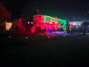 A bus decorated with green and red lights sits outside at night, surrounded by large spider decorations and Halloween props.