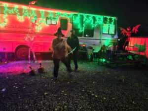 Two people in costumes walk past a Halloween-decorated RV with colorful lights, pumpkins, a skeleton, and a dragon figure in a gravel lot.