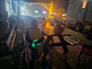 A person in costume sits on a bench at night holding a glowing object; large pumpkins and a plastic container are on the picnic table nearby, with buildings and lights in the background.