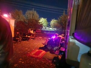 Outdoor campsite at night with a picnic table, chairs, a glowing fire pit, and a red rug on gravel, surrounded by trees and illuminated by colored lights.