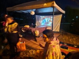People are gathered under a canopy at night with a Halloween movie projected onto a screen, surrounded by carved pumpkins and festive decorations.