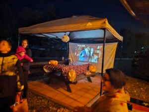 A group of people are near a canopy tent with a picnic table decorated for Halloween; a movie is projected on a screen inside the tent.