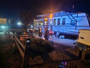 A group of people stands near a lit campfire outside a large white RV at night, with a picnic table and a pickup truck nearby.