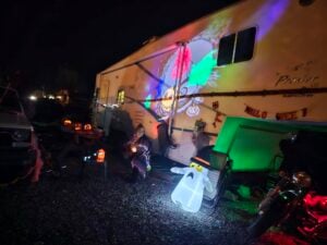 A person sits beside a decorated RV at night with Halloween lights, jack-o'-lanterns, and a ghost decoration. A colorful projection is displayed on the side of the RV.