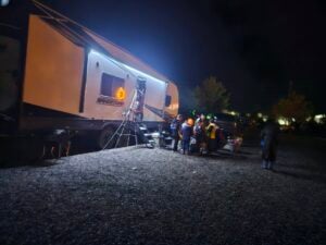 A group of people, including children in costumes, gather outside a lit-up RV at night, possibly for trick-or-treating.