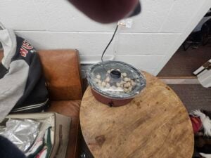 A slow cooker with clear lid sits on a round wooden table, containing meatballs; nearby are a couch with clothing and a small open bag.