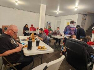 People are seated at tables in a brightly lit room, eating food and talking. Some are facing each other while others are focused on their meals.