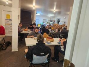A group of people eat and talk around tables in a brightly lit room with posters on the walls and a staircase near the back.