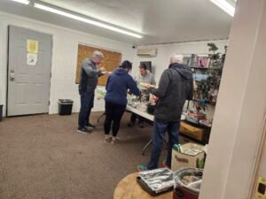Four people serve themselves food from a buffet table inside a room with white walls, a corkboard, and shelves containing books and games.