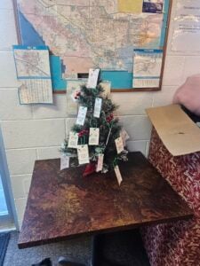 A small decorated Christmas tree with tags hanging from its branches sits on a brown table in front of a map on a wall.