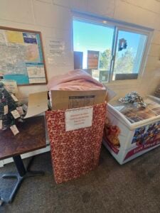 A donation box filled with blankets sits next to a freezer and a small decorated Christmas tree inside a room with a window and maps on the wall.