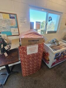 A donation box filled with blankets and bedding stands next to a decorated freezer and a small table inside a community center.