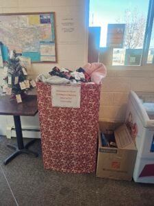A large donation bin filled with clothing and bedding sits next to a cardboard box and a small decorated tree in a community space.