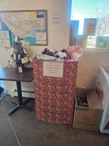 A collection bin filled with donated warm clothing and bedding stands next to a small decorated Christmas tree and cardboard boxes in a community center.