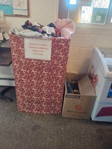 A large red donation bin filled with clothing and bedding sits next to a cardboard box against a wall, under a sign for a community clothing drive.