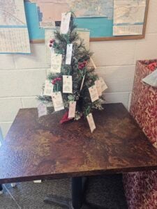Small artificial Christmas tree on a table, decorated with white tags and some red and green ornaments, in front of a wall with maps and papers.