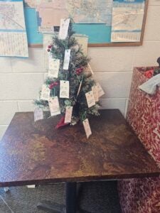 A small decorated Christmas tree sits on a brown table, with tags attached to its branches. A map is visible on the wall behind the tree.