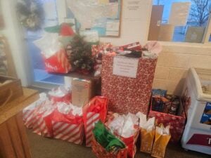 A collection of holiday gift bags and boxes, along with a donation bin labeled "Donations for Women, Children and Housing Items," are arranged near a window and a freezer.
