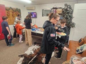 A group of people, including children and adults, stand in line serving themselves food from a counter in a cozy room. A woman in a black jacket with an American flag patch helps a young boy. A television and some decorations are visible in the background.