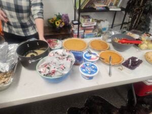 A table filled with various dishes, including multiple pies, a pot of creamy soup, canned drinks, a bowl of mushrooms, and a container of whipped topping. Games and books are visible on a shelf in the background.