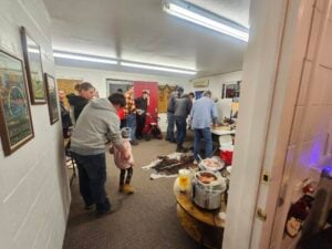 People gathering inside a room with several food dishes set on a table. Some individuals are serving themselves, while others are socializing. The room is decorated with framed pictures on the walls.