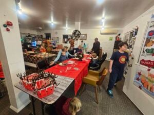 A festive room with a long table covered in a red tablecloth, surrounded by people, including children, engaging in holiday activities. The room is decorated with Christmas-themed items and lights, creating a warm, cheerful atmosphere.