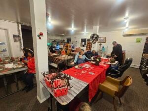 A cozy room with several people seated around tables decorated with red tablecloths and holiday decorations. Shelves with various items are in the background. It appears to be a festive gathering or celebration.
