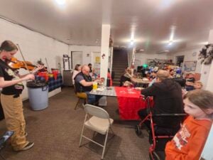 A group of people gather in a community room. A person plays the violin on the left while others sit around tables covered with tablecloths. Some are engaged in conversation, and there's a young person observing the scene in the foreground.