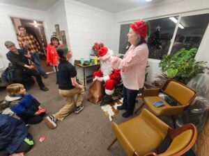 A festive scene with Santa sitting indoors, handing something to a child kneeling nearby, while people, including a woman in an elf hat, are smiling and watching. Holiday decorations and chairs are in the room.