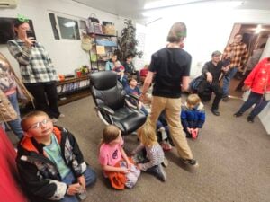 A group of children and adults are gathered in a room with shelves of books and decorations. Some children are sitting on the floor, while others are standing or sitting in chairs. An adult takes a photo with a smartphone.