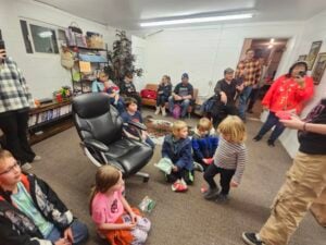 A group of children and adults gather in a cozy, cluttered room with white walls. Some are seated on chairs and couches, while others stand. The room is decorated with bookshelves and plants, creating a casual, lively atmosphere.