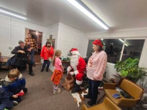 A group of people, including children, gather around a person dressed as Santa Claus in a festive room. A girl talks to Santa while others observe. The room is decorated with a Christmas tree and holiday decorations.