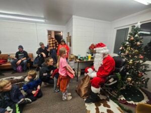 A group of children and adults gather around a person dressed as Santa Claus in a white-walled room. Santa is seated next to a decorated Christmas tree. Some kids are holding gifts and talking to Santa. The atmosphere is festive and cheerful.