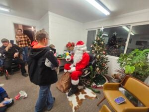 A child stands in front of Santa Claus, who is seated and holding a gift in a room decorated with a Christmas tree. Several people are watching, and festive ornaments can be seen around the room.