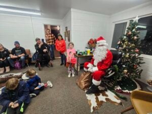 A child stands next to Santa Claus in a room decorated with a Christmas tree. Adults and children are seated on chairs, watching the scene. Some people hold snacks or drinks, and festive decorations can be seen throughout the room.