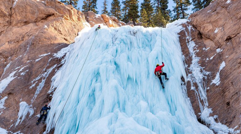 Ice climber ascending a tall frozen waterfall with a belayer below, surrounded by generic canyon walls and snow-dusted evergreens, in bright winter daylight.