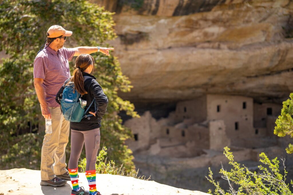 An adult and a child stand on a ledge near Grand Junction, admiring ancient cliff dwellings built into a rocky alcove. The adult points ahead while the child, in rainbow-striped socks and a blue backpack, takes in the Colorado scenery framed by trees.