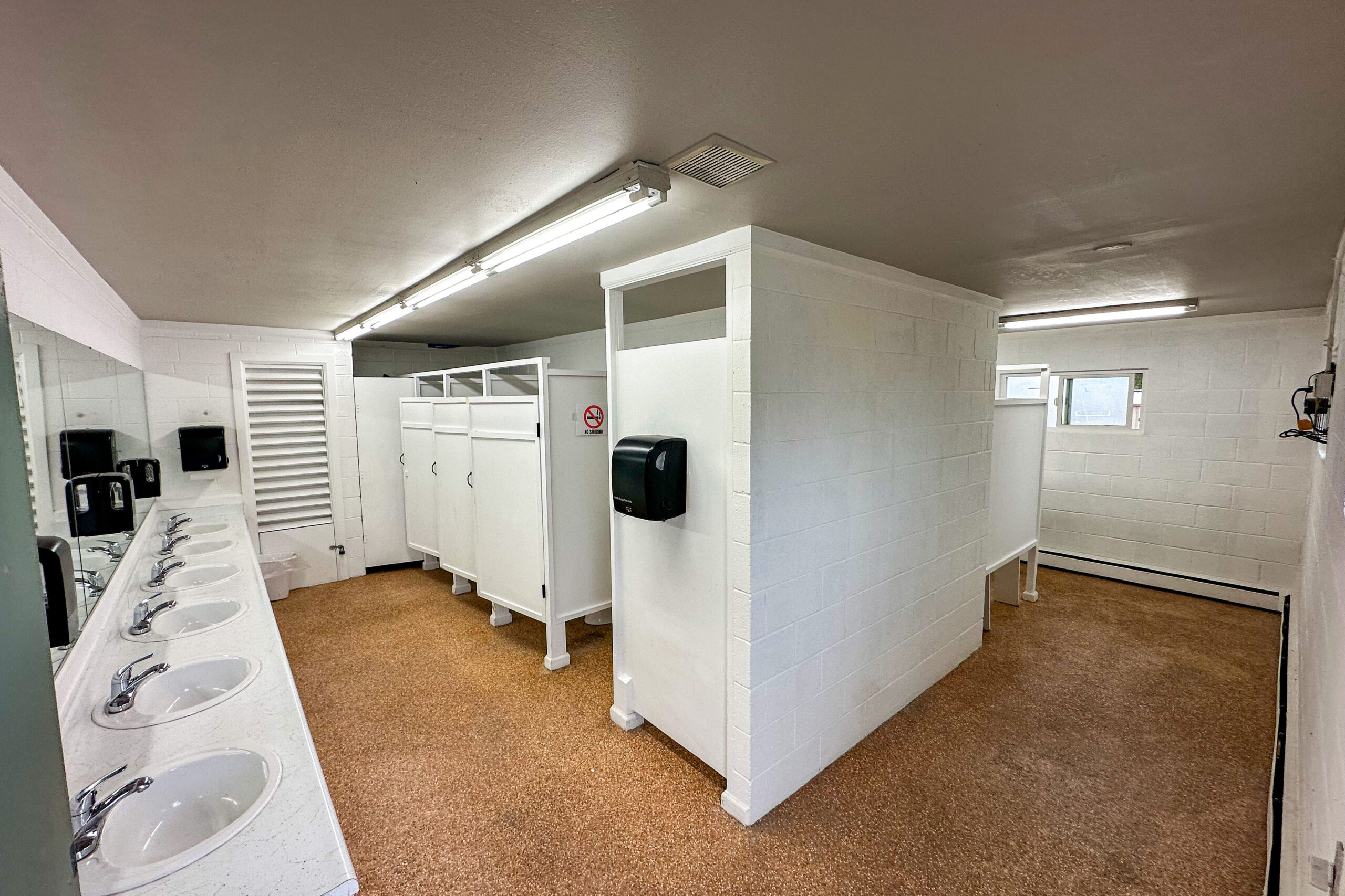 A clean public restroom with five sinks, mirrors, soap dispensers, paper towel dispensers, and several toilet stalls with doors. The floor is brown and the walls are white.
