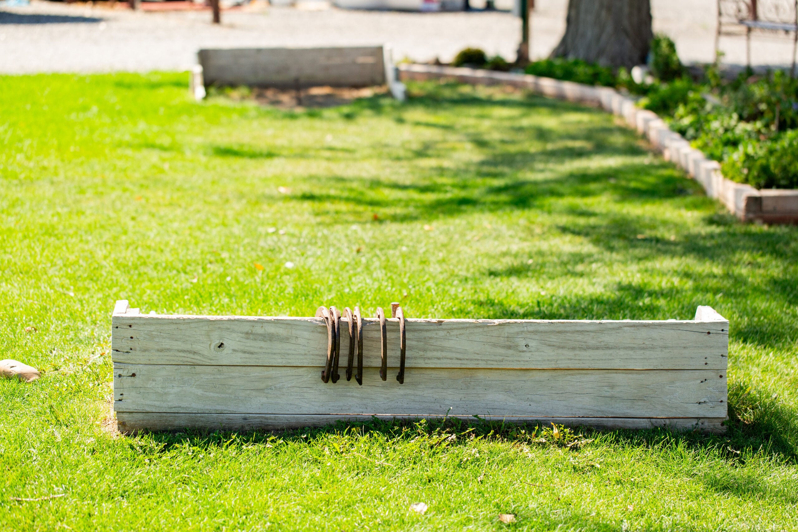 A wooden horseshoe pit with four horseshoes hanging on the front, set on green grass with another pit in the background.