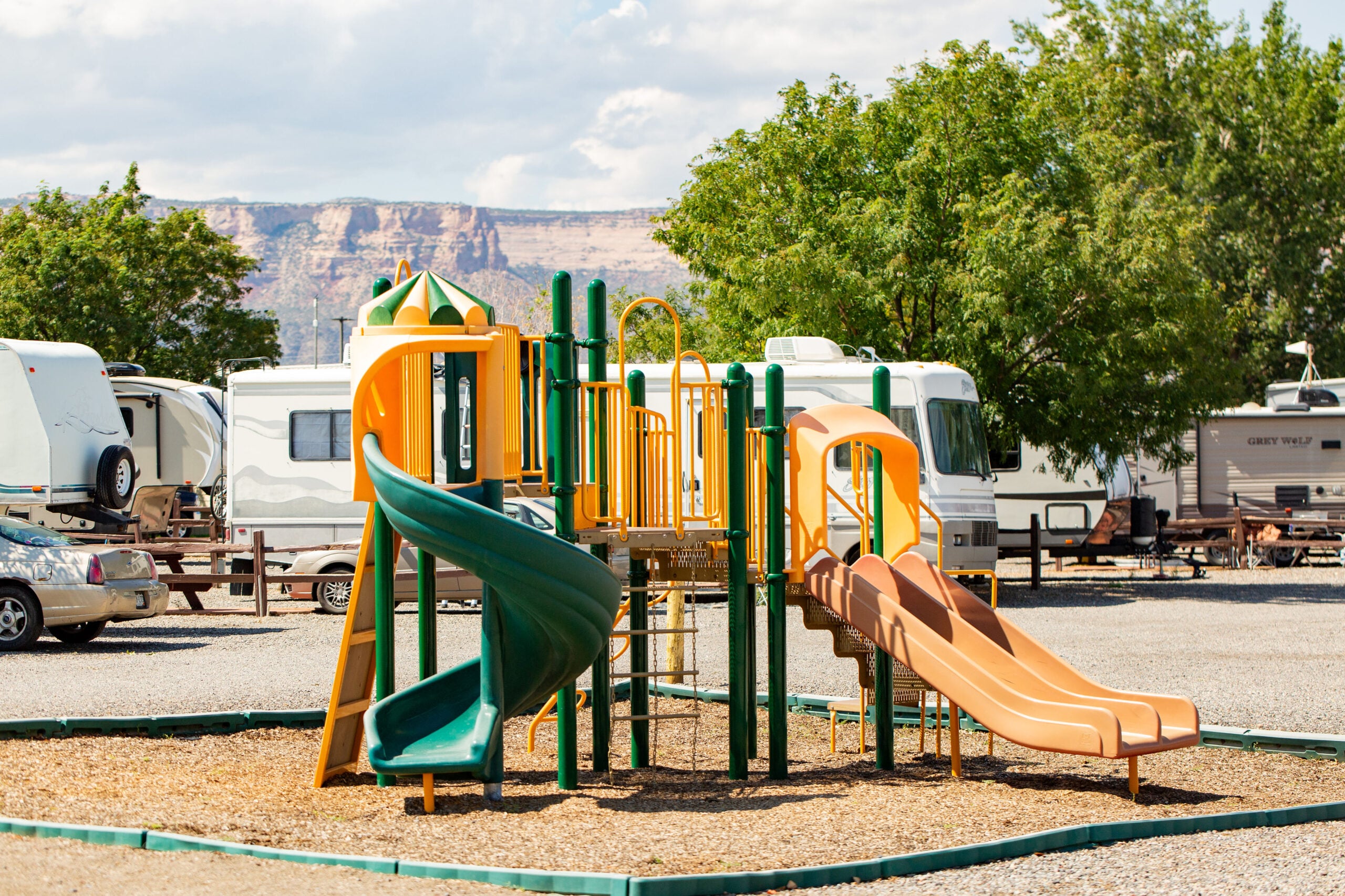 Colorful playground with slides and climbing structures in a gravel area, surrounded by RVs and trees, with mountains visible in the background.