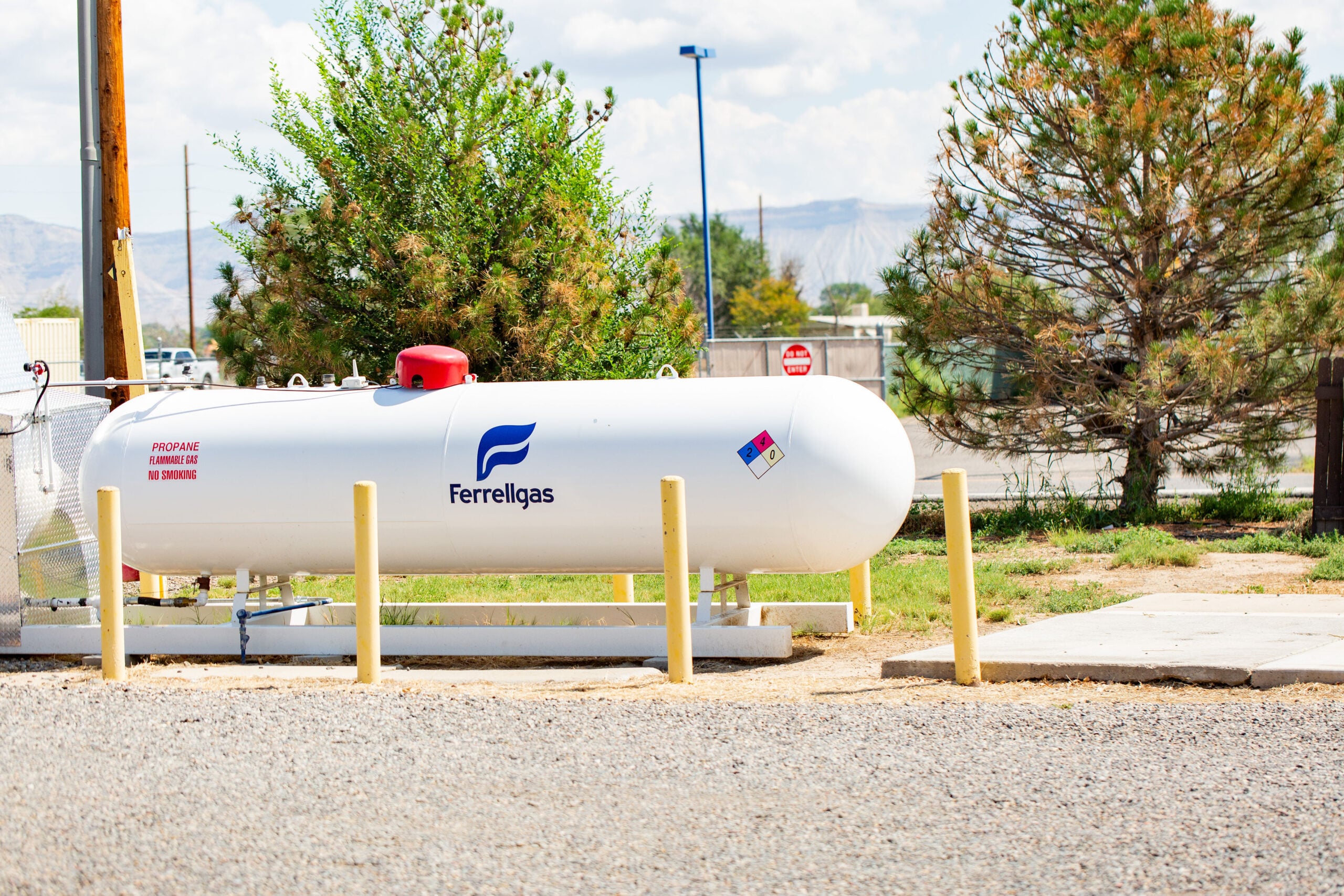 A large white Ferrellgas propane tank is situated outdoors, surrounded by yellow posts, with trees, a fence, and a street in the background.