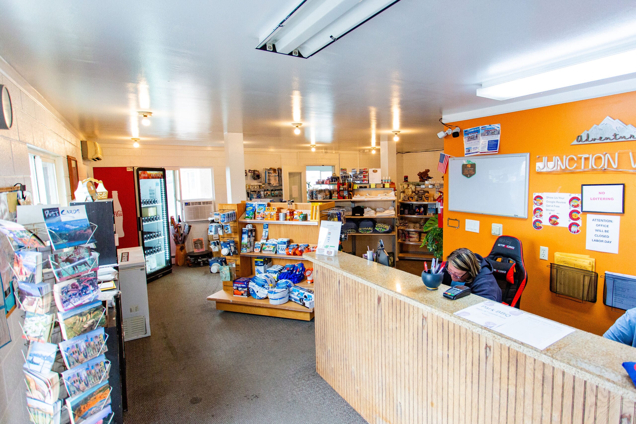 A small convenience store and reception area with shelves of snacks and supplies, a brochure stand, and a person working behind a counter.