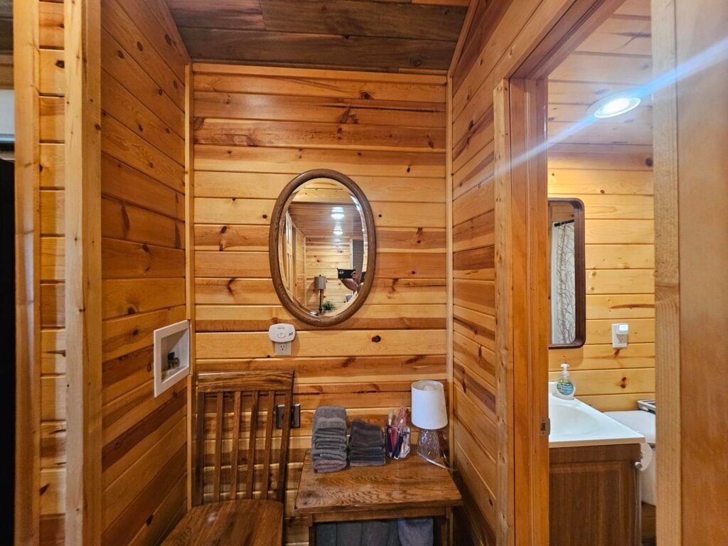 Small wooden room with a mirror reflecting the sink area, a bench with folded towels, lamp, and toiletries beneath it. Walls and ceiling are made of light-colored wood panels.
