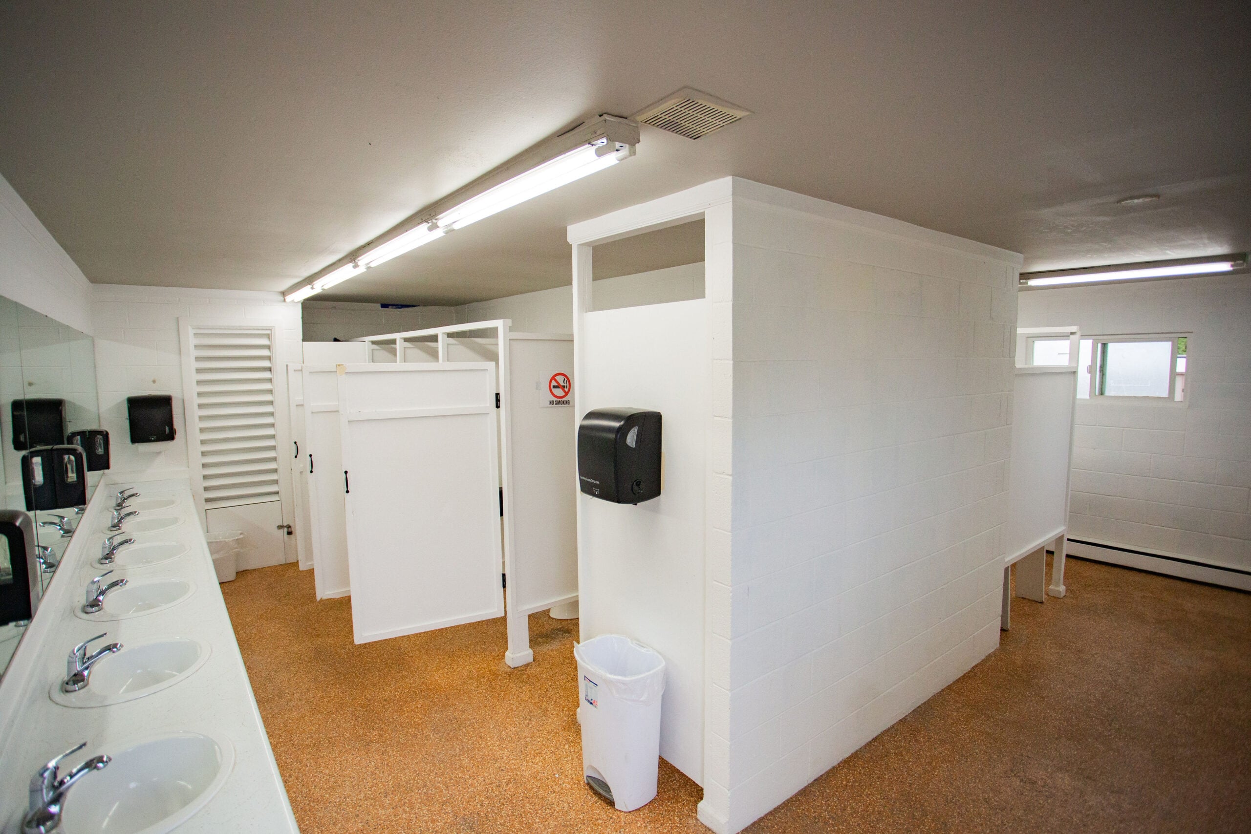 A clean public restroom with white stalls, sinks along one wall, soap dispensers, a paper towel dispenser, and a trash can on a tan floor.