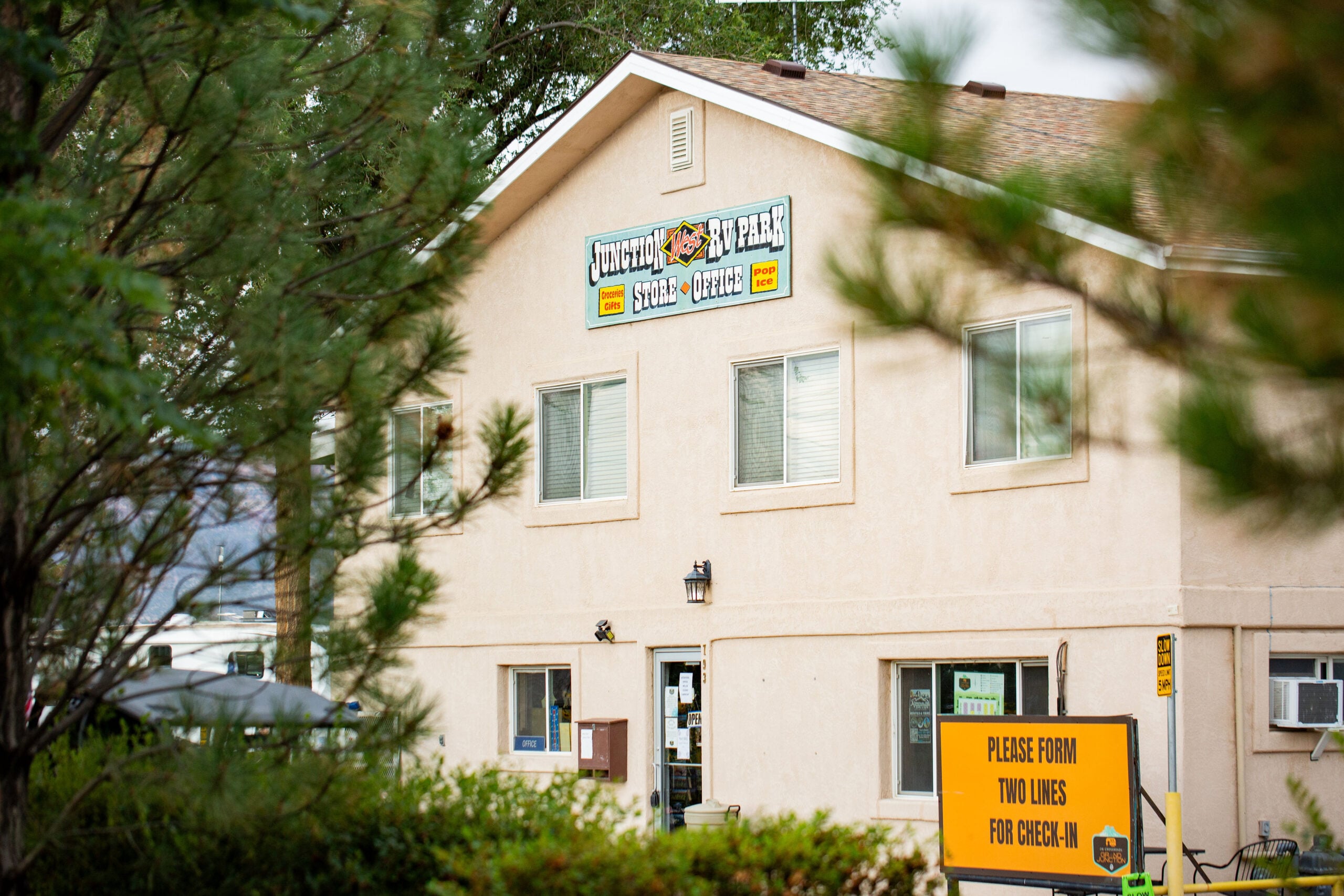 A beige two-story building with a sign reading “Lighthouse RV Park,” partly obscured by trees; a yellow check-in instruction sign is visible in the foreground.